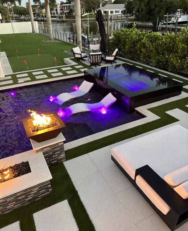 This striking pool design combines sleek, contemporary elements with luxurious details to create a unique backyard oasis. The centerpiece is a pristine infinity edge pool with reflective black tiles, producing a stunning visual effect as water gently cascades over the edges. Integrated fire bowls, positioned at each corner of the pool, add a dramatic touch and provide ambient warmth, perfect for evening gatherings. The pool is surrounded by meticulously manicured artificial turf and stylish white pavers, leading to comfortable poolside seating areas and a basketball hoop for fun and relaxation. This pool is not only a place to swim but a modern retreat designed for both entertainment and tranquility.
